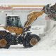 Excavator piling snow in a parking lot beside a gas station.